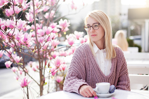 Young woman is drinking coffee in cafe on spring city streets. Attractive girl is enjoying of magnolia garden at sunset outside. Blooming bushes of flowers are pink and purple.