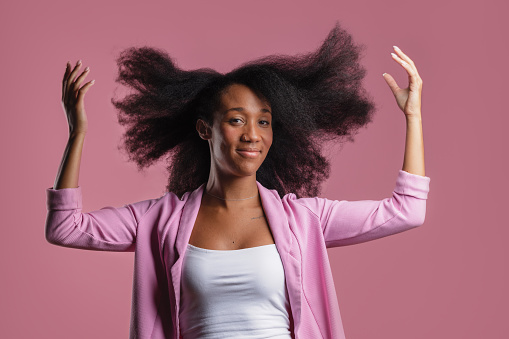 Portrait of a cheerful young woman in casual clothing raising her arms with confidence against a pink background.