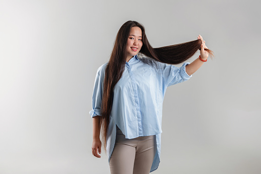 A young adult woman in smart casual attire holds and showcases her long hair with a light smile, isolated on a neutral background.