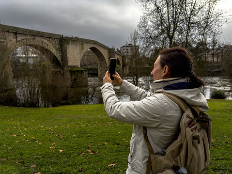 A solo traveler, clad in a white jacket, stands in a verdant park, capturing the serene beauty of an ancient stone bridge over a tranquil river with her smartphone. The contrast of historical architecture and natural scenery encapsulates the essence of solo travel - immersing oneself in the environment and preserving memories of wanderlust.