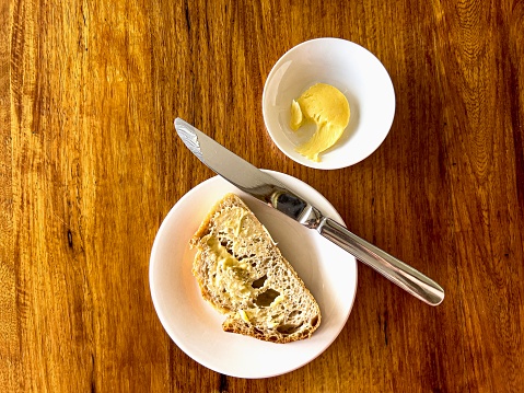 Horizontal high angle closeup photo of a slice of freshly baked, buttered sourdough bread on a round white ceramic plate with a butter knife and a round of cultured butter on a small round white side bowl on a wooden table in a fine dining restaurant. Milton, south coast NSW.