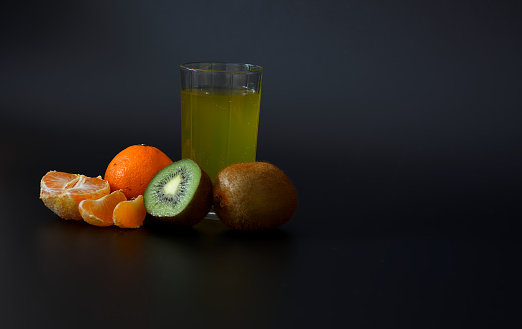 A tall glass of freshly squeezed fruit juice on a black background, next to tangerine slices and a kiwi fruit. Close-up.