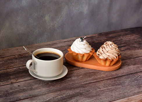 Two basket cakes with cream and a cup of black coffee on a saucer with a spoon on a wooden table. Close-up.