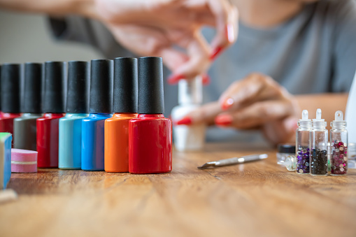 Colored nail polishes with a background of a woman painting her nails on a table with many accessories for work.