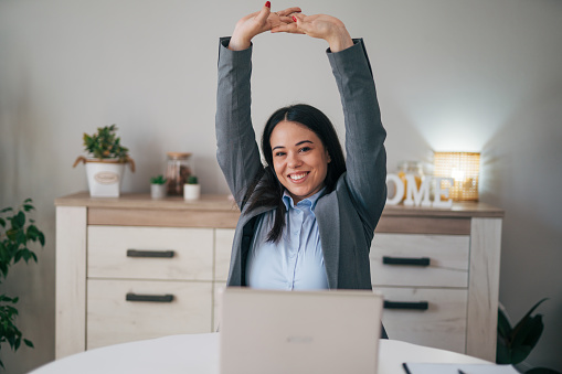 Happy relaxed young woman sitting in her dining room with a laptop in front of her stretching her arms above her head