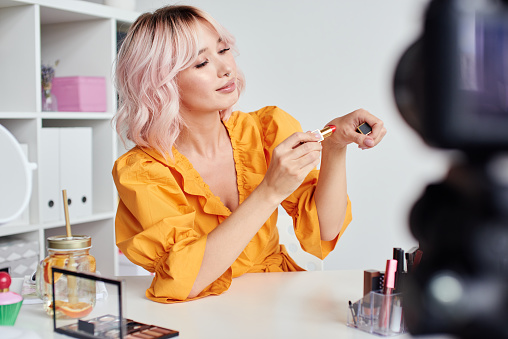 Young beautiful blond asian woman is recording make up tutorial video for beauty blog, wearing terracota blouse. Video blogging, isolation, stay home concept