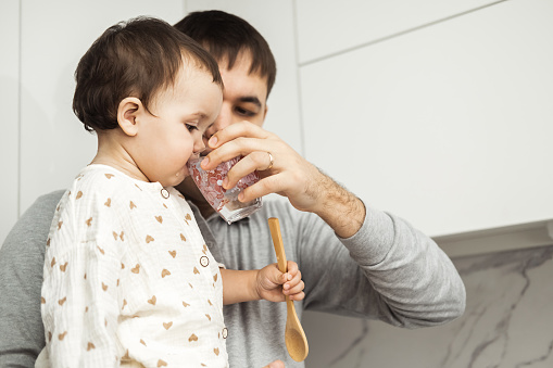 A caring father stands in the kitchen with a daughter in his arms, singing his baby with water from a glass