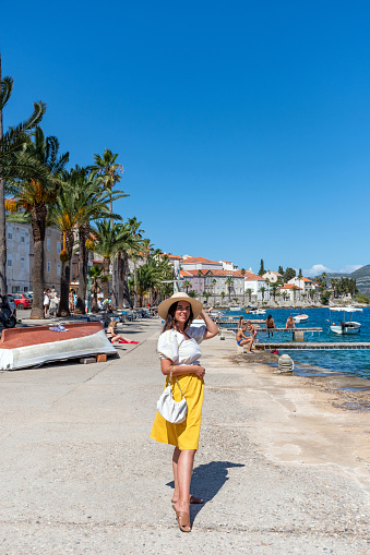 A woman in stylish summer clothes at the seafront with piers, palm trees, and idyllic houses in Korcula town, Croatia.