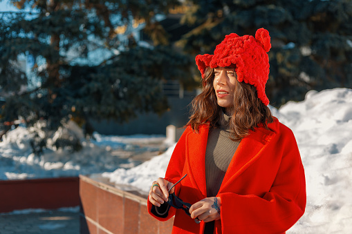 Brunette woman in a red hat with ears and a red coat on a snowy background holding a sunglasses in her hand