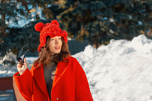 Woman in a red hat with ears and a coat against a background of snow holding sunglasses in her hand