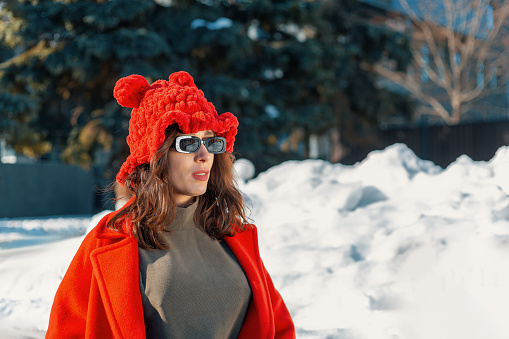 Brunette in an interesting hat with sunglasses and a red coat standing on a winter background