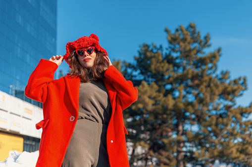 Brunette in a red coat and hat standing against the background of a clear sky and a green tree