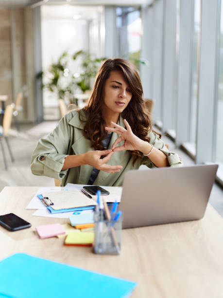 Deaf businesswoman having a video call over laptop in the office. Young female entrepreneur using sign language during an online business meeting over a computer in the office. sign language stock pictures, royalty-free photos & images