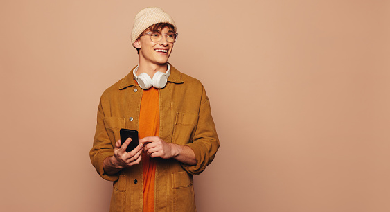 Happy man, wearing casual denim clothing, stands against a peach background in a studio. He holds a smartphone, using a mobile app and texting. Vibrant and colorful, this modern image portrays a cheerful and tech-savvy individual.