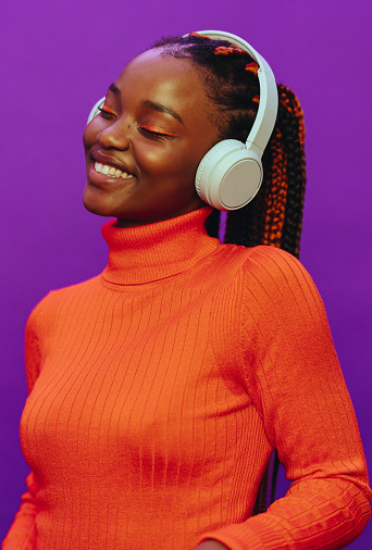Happy woman with colorful two-tone braids. She stands against a vibrant purple background, wearing casual clothing and headphones. Her modern makeup and trendy eyeliner complement her cheerful expression as she enjoys streaming music.