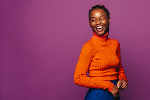 Happy young woman in her 20s stands in a studio, wearing casual clothing and vibrant purple polo neck. Her stylish two-tone braids highlight her fashionable look, showcasing her cheerful smile and trendy makeup.