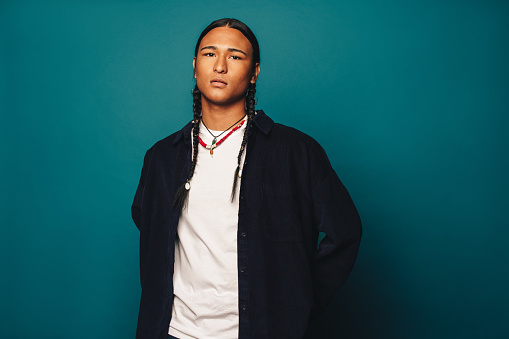 This young man exudes self-confidence standing in the studio. With long braided hair and stylish jewelry, including a necklace, he exudes ethnic pride. Casual clothing and a blue background further enhance his portrait.