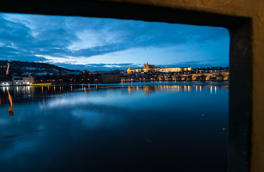 Prague sprawling city at night, its buildings mirrored in the calm waters of the river Vltava as a lone bridge stretches across the vast landscape under a cloudy sky