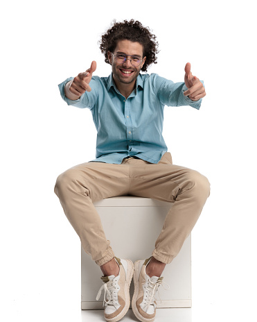 joyful casual man sitting on a chair on white background and making thumbs up sign