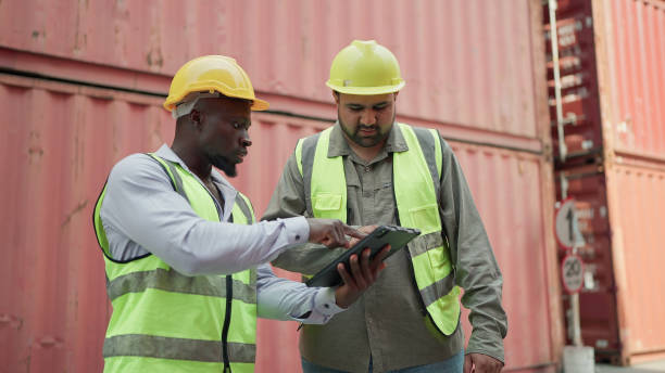 istock Two male container engineers working on tablet inspecting containers around a shipping yard of logistic freight forwarder company. Two engineer checking container storage with cargo container 1951942682