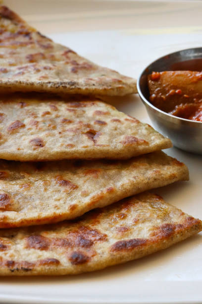 Stock photo showing close-up view of a breakfast platter of triangular slices of aloo parathas served on a white plate with mango chutney pickle, traditional Indian recipe. Savoury bread stuffed with garam masala potatoes and onions. imagen de fotograma completo del buffet de platos de desayuno indios, rebanadas triangulares de aloo parathas en un plato blanco, pan salado relleno de papas garam masala, molde de metal de pepinillo de chutney de mango, vista elevada, enfoque en primer pl