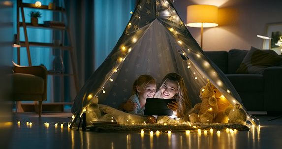 Mom, daughter and home tent with tablet, night and lights on living room floor. Happy, internet and streaming movies together for holiday, technology and home with digital device, watching and online