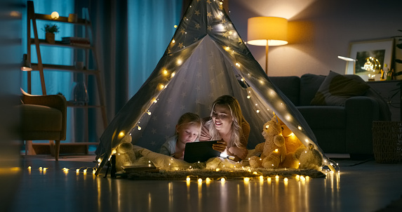 Child, fort and mother with tablet in a family home at night with sleepover, movie or youth game. Lights, happy and young girl with a mom together with tech, series streaming and website in a bedroom