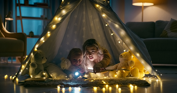 Mom, daughter and tent with torch, night and lights on living room floor, searching for monsters. Looking, house and pointing for playing games, child and parent for childhood memories and holidays