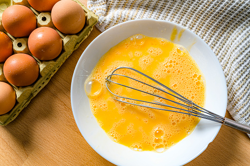 Overhead shot of scrambled raw eggs in a white bowl. A wire whisk and an eggs carton complete the composition. High resolution 42Mp outdoors digital capture taken with SONY A7rII and Zeiss Batis 25mm F2.0 lens