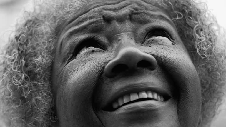 One black senior woman gazing up at sky close-up face in monochromatic, black and white. One African American elderly person in 80s looking up at sky with GRATITUDE