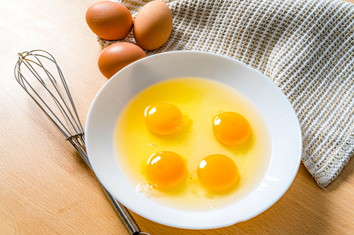 Four raw eggs in a white bowl. A wire whisk and whole eggs complete the composition. High resolution 42Mp outdoors digital capture taken with SONY A7rII and Zeiss Batis 25mm F2.0 lens