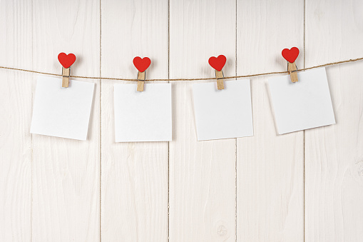 Clothespins with red hearts and empty sheets of paper on string against white wooden background. Valentines day holiday, romantic concept. Mockup, template, copy space