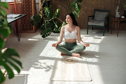 Content female practitioner peacefully performing padmasana in lotus position on cozy yoga rug. Charming woman sitting on floor of stylish exotic room and lloking to the window