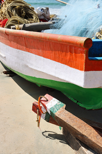 Stock photo showing close-up view of a wooden fishing boat hauled up on sand besides rustic wood blocks for mooring with view of sea and breaking waves at low tide. The fishing boat, full of ropes and fishing net, and wood chocks are painted with  saffron (orange), white and green stripes like the National flag of India (tricolour).