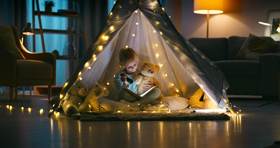 Night, teddy bear and girl with a book, relax and happy with education, fairy lights and hobby. Kid reading a story, stuffed animal and child with a torch, playful or home with education, tent or joy