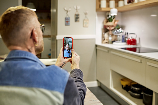 Rear view of mature man sitting in domestic kitchen, focus on smart phone display and partner conversing from his home.