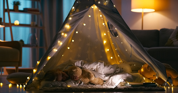 Child, fort and sleeping with blanket in a family home at night with sleepover, nap and rest. Lights, teddy and young girl in a lounge with kid, teepee and evening in a living room in a house