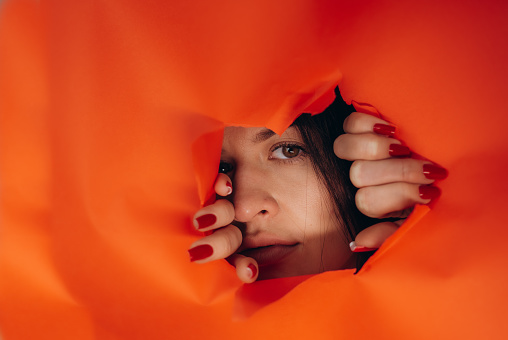 Surprise. Portrait of a young caucasian woman looking through the hole in orange paper. Distrustful look. Women's curiosity and gossip. A jealous wife. Copy space.