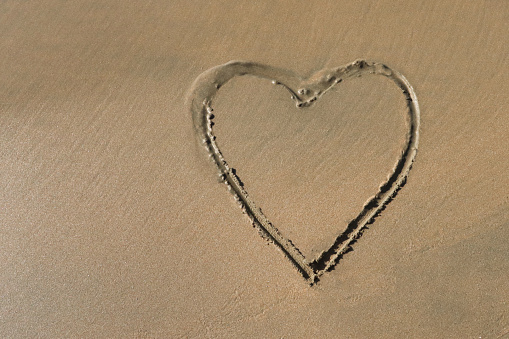 Stock photo showing heart shape drawn on sunny beach in sand with stick in soft, golden, wet sand on seaside coastline.