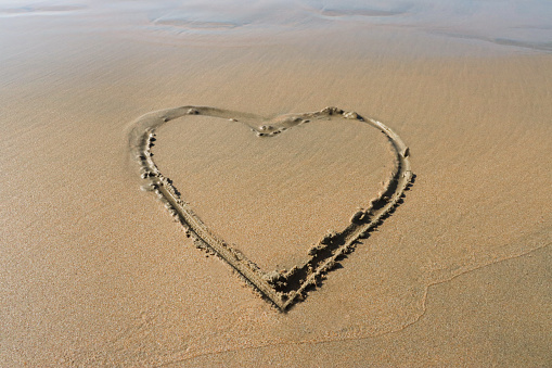 Stock photo showing heart shape drawn on sunny beach in sand with stick in soft, golden, wet sand on seaside coastline.