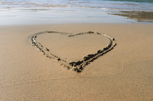 Stock photo showing heart shape drawn on sunny beach in sand with stick in soft, golden, wet sand on seaside coastline.