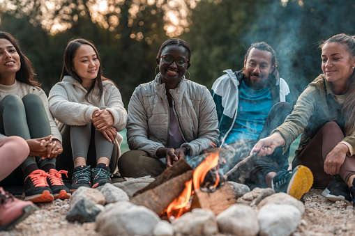 Mixed race friends come together for a delightful day in nature, basking in the warmth of a bonfire, fostering connections and savoring the simple pleasures of shared company.