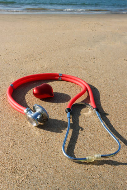 istock Close-up image of model, plastic red heart by stethoscope on sunny sandy beach, waves breaking at low tide, sea and coastline, stress free health and wellbeing concept, focus on foreground, copy space 1952032566