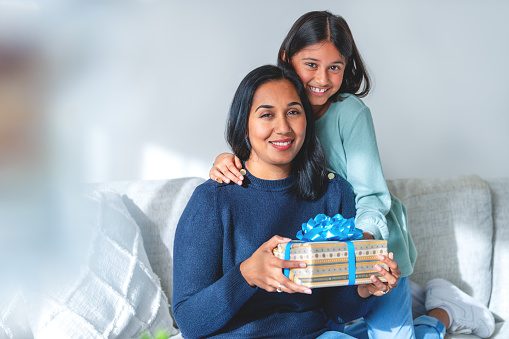 Loving daughter giving her mother a gift on the sofa at home. Could be a Mothers Day gift, or a birthday present. Both are happy and smiling looking at camera