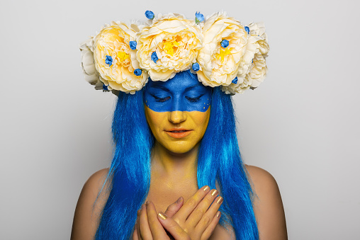 Young woman with yellow blue face art, wreath of peonies on head, holding hands to chest, praying for salvation on isolated gray background. Creative concept for Flag Day in Ukraine. Portrait close-up