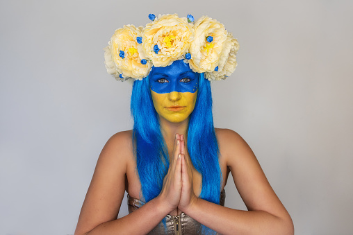 Young woman with yellow blue face art, wreath of peonies on head, praying for salvation on an isolated gray background. Creative concept for Flag Day in Ukraine. Copy space. Girl looking at camera