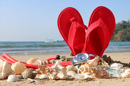 Stock photo showing close-up view of a stethoscope on top of a pile of seashells in front of a pair of red flip flops standing up, forming a heart shape, in the sand on a sunny, golden beach with sea at low tide in the background. Stress free health and wellbeing concept.