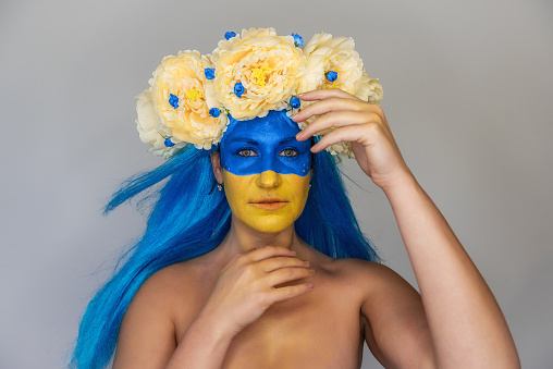 Creative close-up portrait of young Ukrainian patriot woman with yellow blue face art, wreath of flowers on head on isolated gray background. Flag Day, Independence Day in Ukraine. Copy space