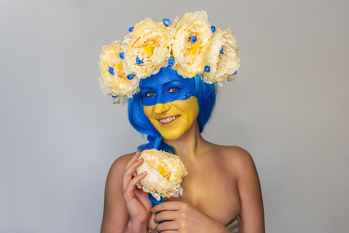 Creative close-up portrait of young happy Ukrainian patriotic woman with yellow blue face art, wreath of flowers on head, holding braid in hands on isolated gray background. Flag Day, Independence Day