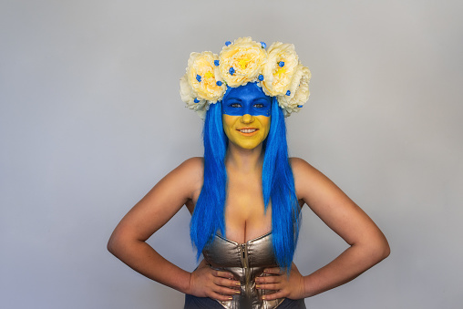 Beautiful creative young woman smiling while looking at camera on isolated background. Girl dressed in bright corset, blue hair, yellow blue face art, wind blows hair, wreath of spring flowers on head